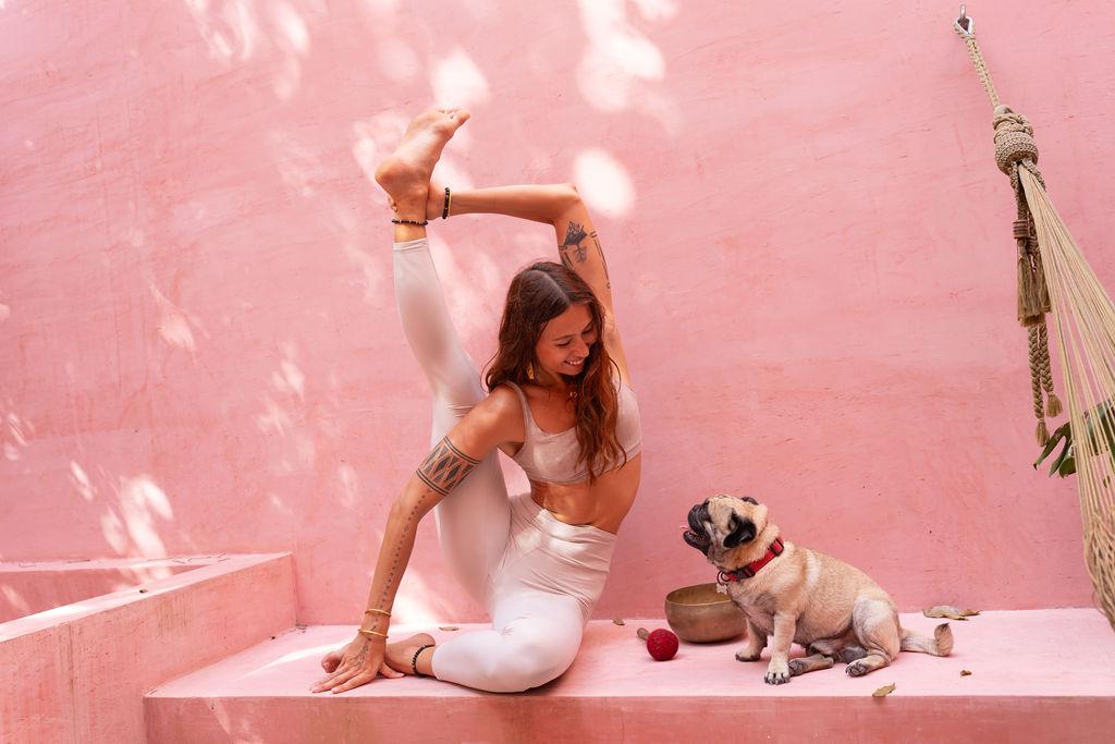 Natalia Orlova practising a standing balance against a pink wall in Tulum, her pug sitting in the foreground